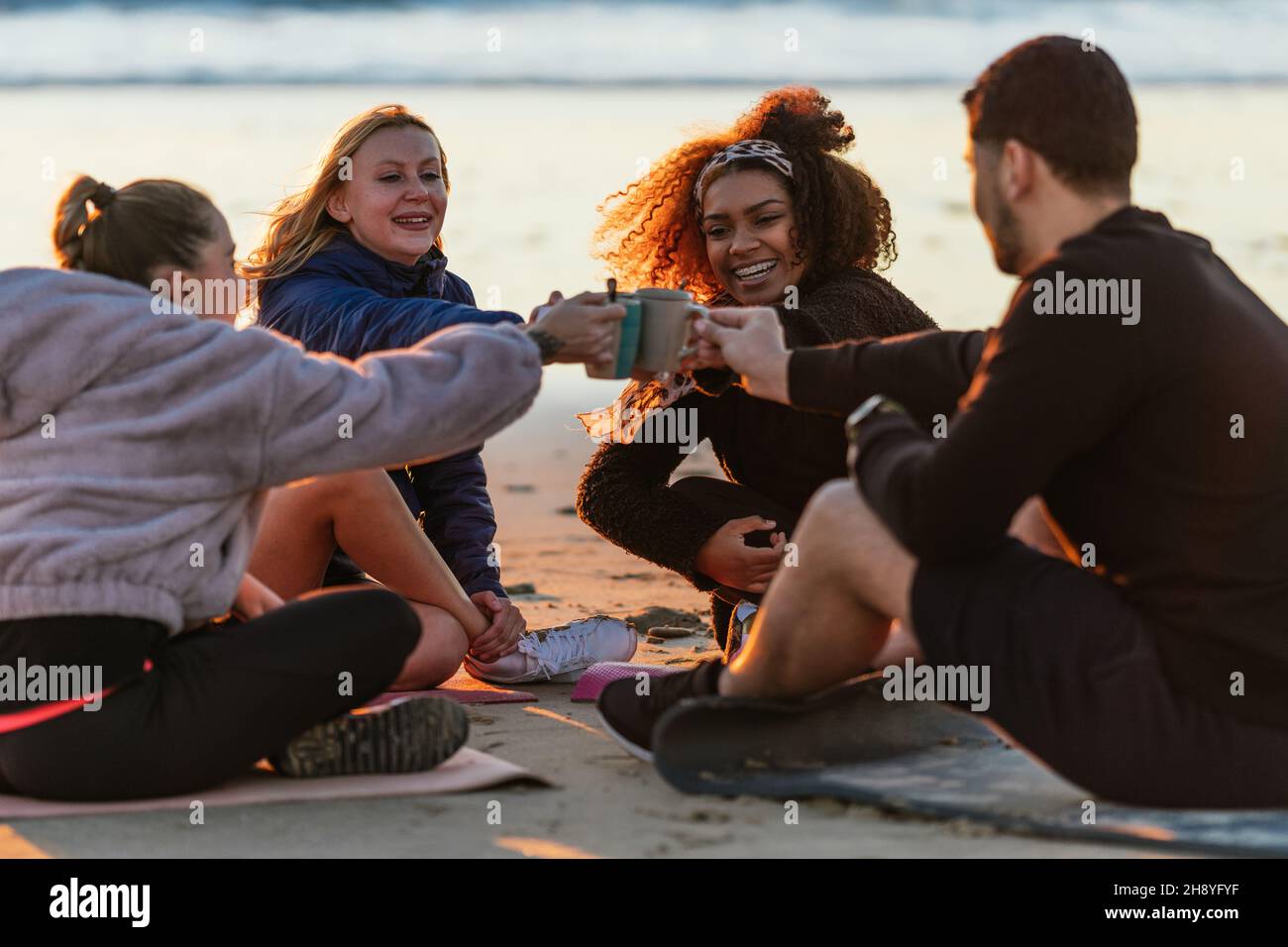Eine multiethnische Gruppe von Menschen, die während des Sonnenuntergangs mit Tee am Strand toasten Stockfoto