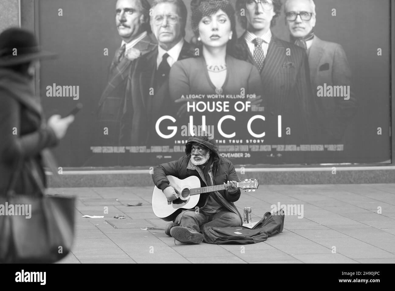London, England, Großbritannien. Älterer schwarzer Mann, der auf dem Leicester Square vor einem Filmplakat für das Haus Gucci anschellt/bettelt - Dezember 2021 Stockfoto