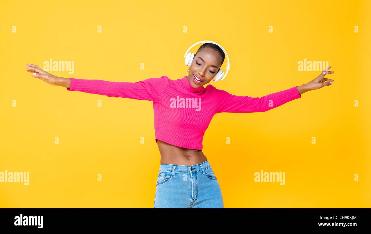 Porträt einer glücklichen afroamerikanischen Frau, die Kopfhörer trägt und Musik hört und auf einem farbenprächtigen, gelben, isolierten Hintergrund tanzt Stockfoto