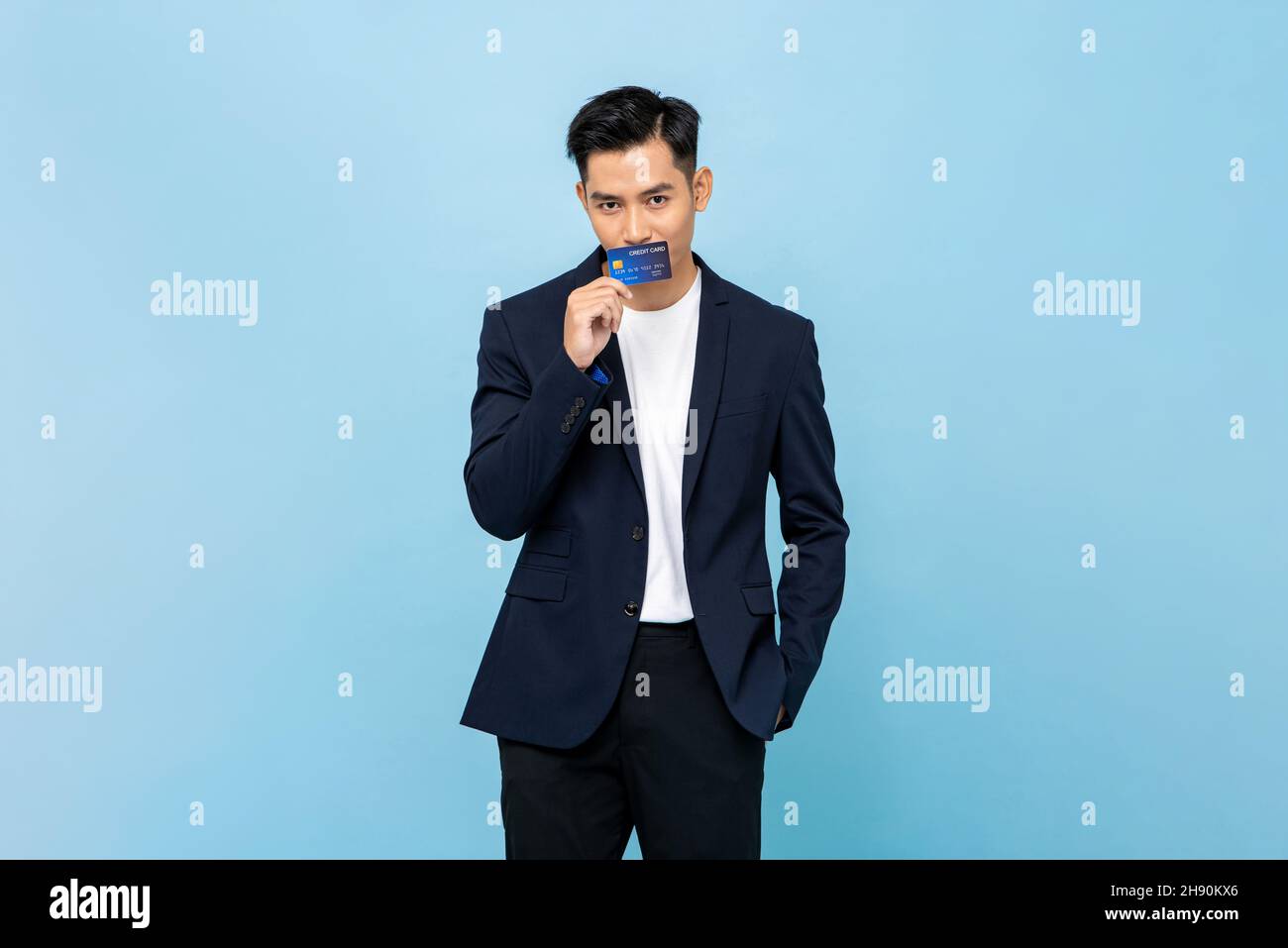 Junger hübscher asiatischer Mann im halbformalen Blazer küsst Kreditkarte in hellblau isoliert Studio Hintergrund Stockfoto