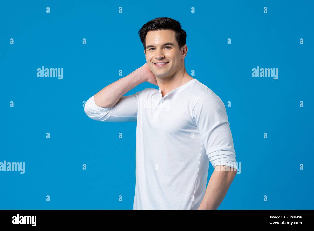 Waist-up-Aufnahme von lächelndem glücklichen jungen, hübschen kaukasischen Mann in schlichtem weißen T-Shirt, der auf einem isolierten blauen Hintergrund die Kamera anschaut Stockfoto