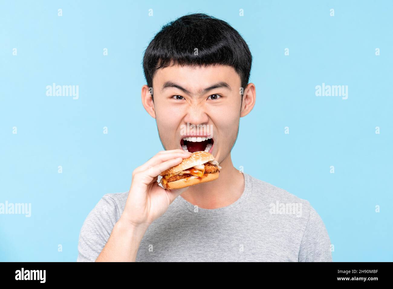 Junger hungriger asiatischer Mann, der Hamburger im isolierten hellblauen Studiohintergrund isst Stockfoto