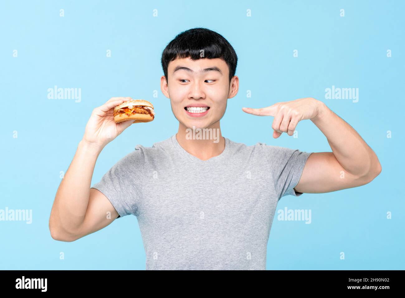 Junger asiatischer Mann zeigt und schaut auf einen Hamburger in hellblauem, isoliertem Hintergrund Stockfoto