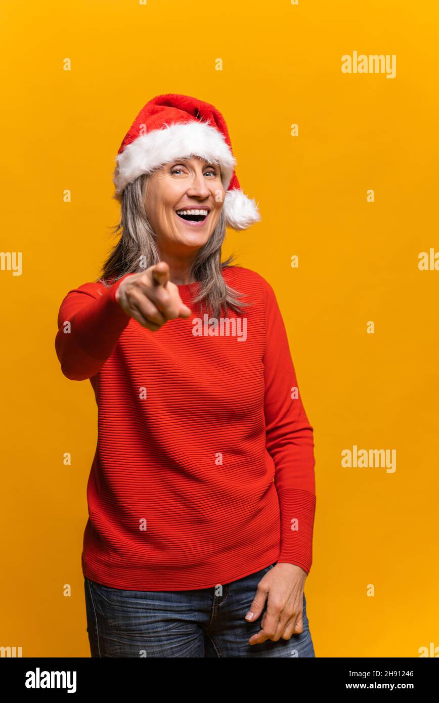 Fröhliche glückliche ältere Frau in santa hat zeigt Zeigefinger auf die Kamera und lacht, vertikales Porträt mittleren Alters Frau, die Sie wählt. Werbekonzept für das neue Jahr Stockfoto
