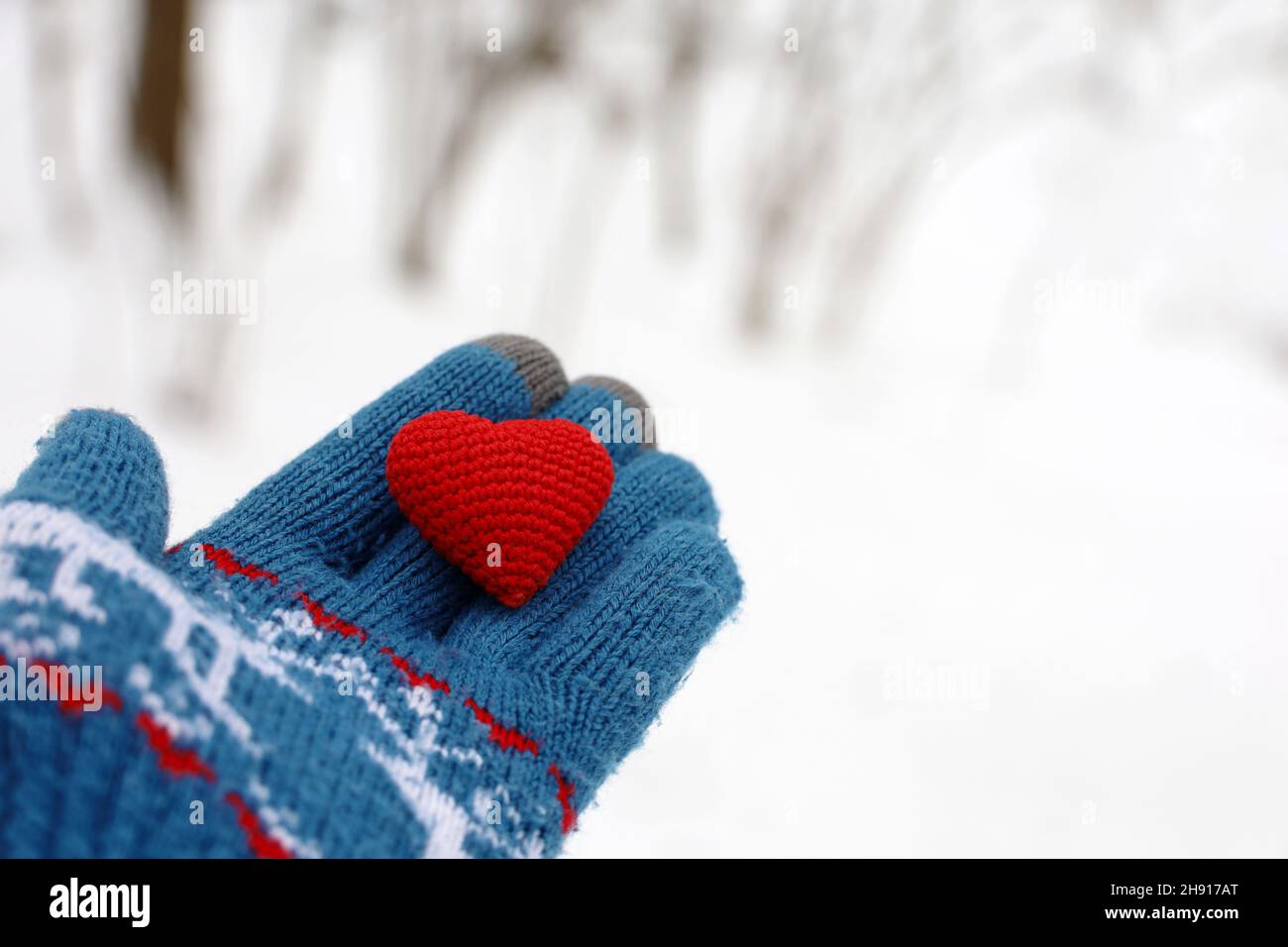 Rotes Herz auf Handfläche in warmem Strickhandschuh gegen den Winterpark und Schnee. Konzept einer romantischen Liebe, Valentinstag oder Nächstenliebe Stockfoto