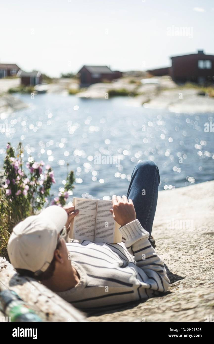 Mann liest Buch, während er sich an sonnigen Tagen auf einem Felsen legt Stockfoto