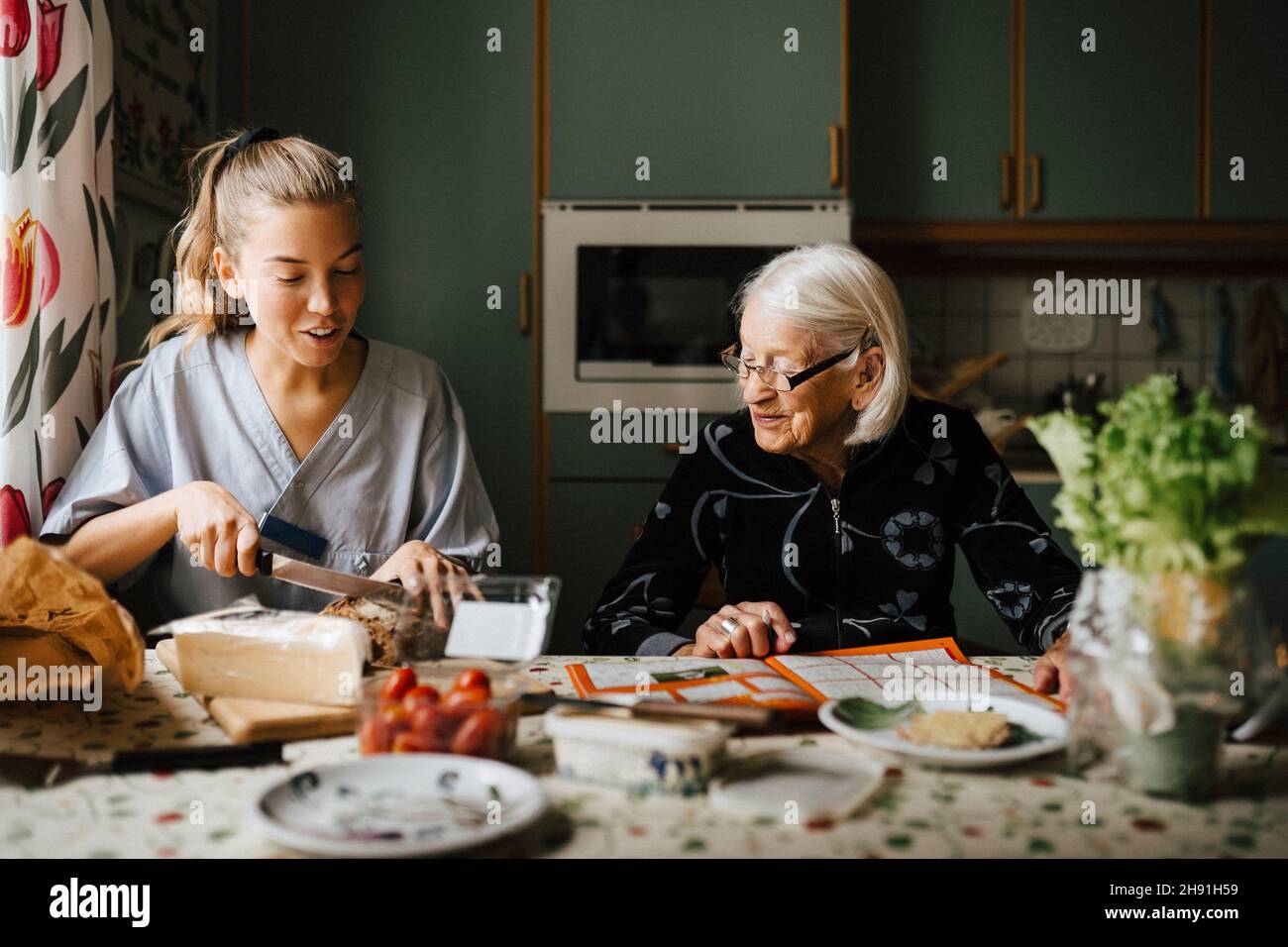 Die Betreuerin schneidet Brotlaib am Esstisch, während sie von einer älteren Frau in der Küche sitzt Stockfoto