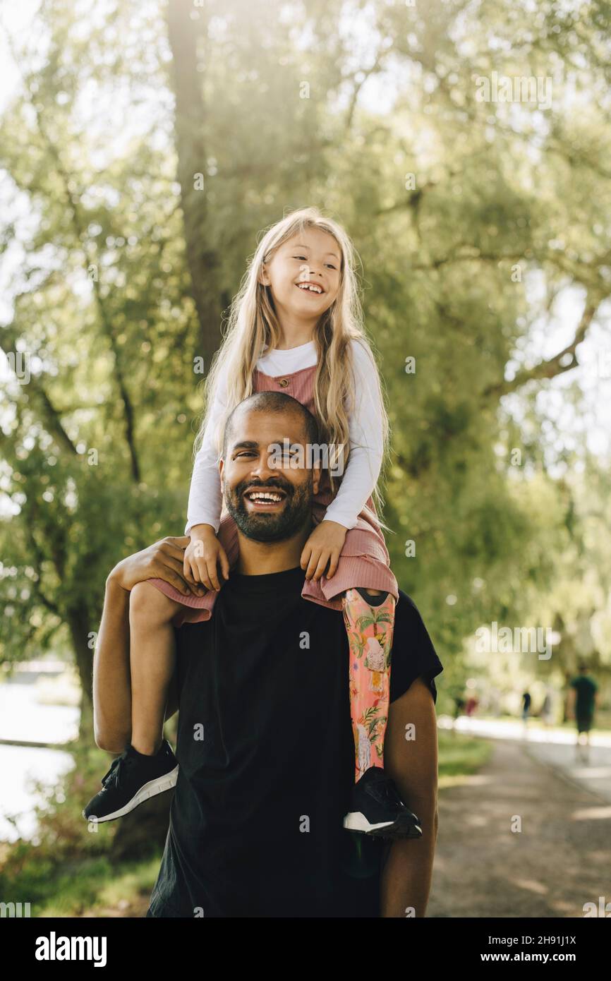 Fröhlicher Vater, der eine behinderte Tochter auf den Schultern trägt, während er im Park spazieren geht Stockfoto