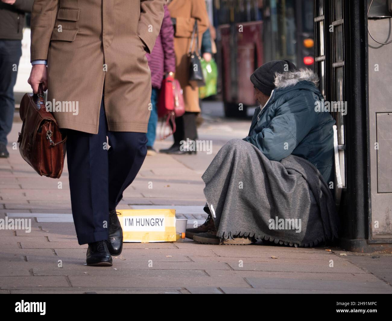 Reiche Arme teilen die Ungleichheit, männlicher Bettler mit dem Schild „Ich bin hungrig“, bettelt in Mayfair London, während wohlhabende, gut gekleidete Menschen an ihnen vorbei gehen Stockfoto