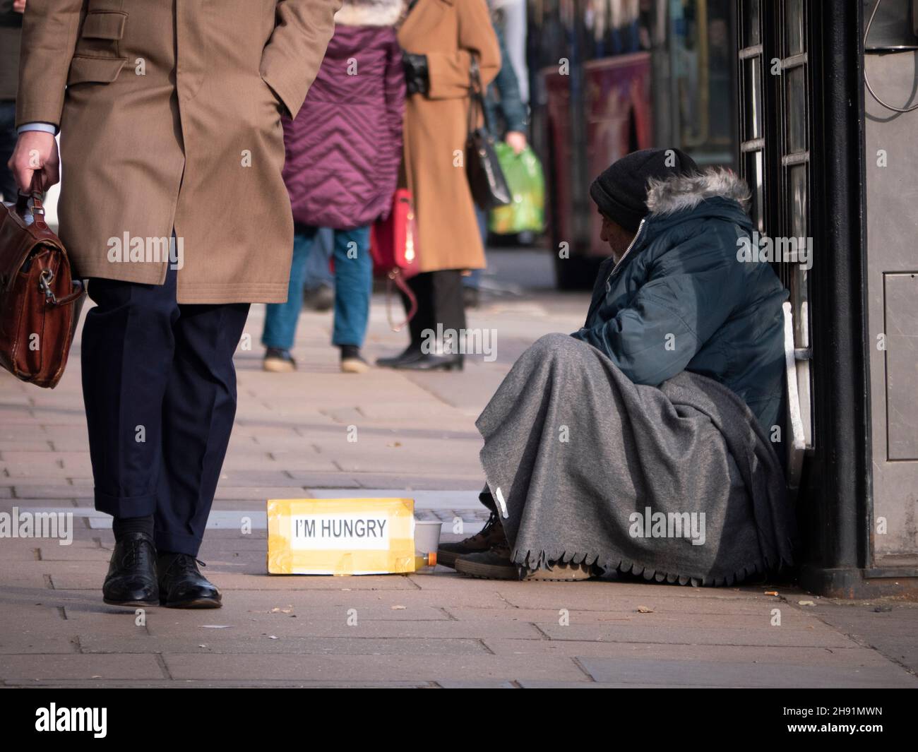 Reiche Arme teilen die Ungleichheit, männlicher Bettler mit dem Schild „Ich bin hungrig“, bettelt in Mayfair London, während wohlhabende, gut gekleidete Menschen an ihnen vorbei gehen Stockfoto