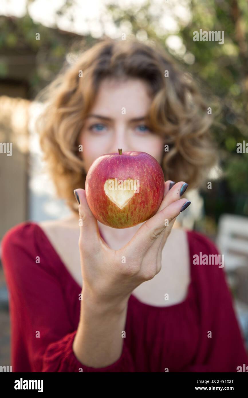 Schöne junge Frau hält einen roten Apfel mit einem Herz in ihm vor ihrem Gesicht geschnitzt. Selektiver Fokus auf den Apfel. Richtige Vitaminernährung. Surpr Stockfoto