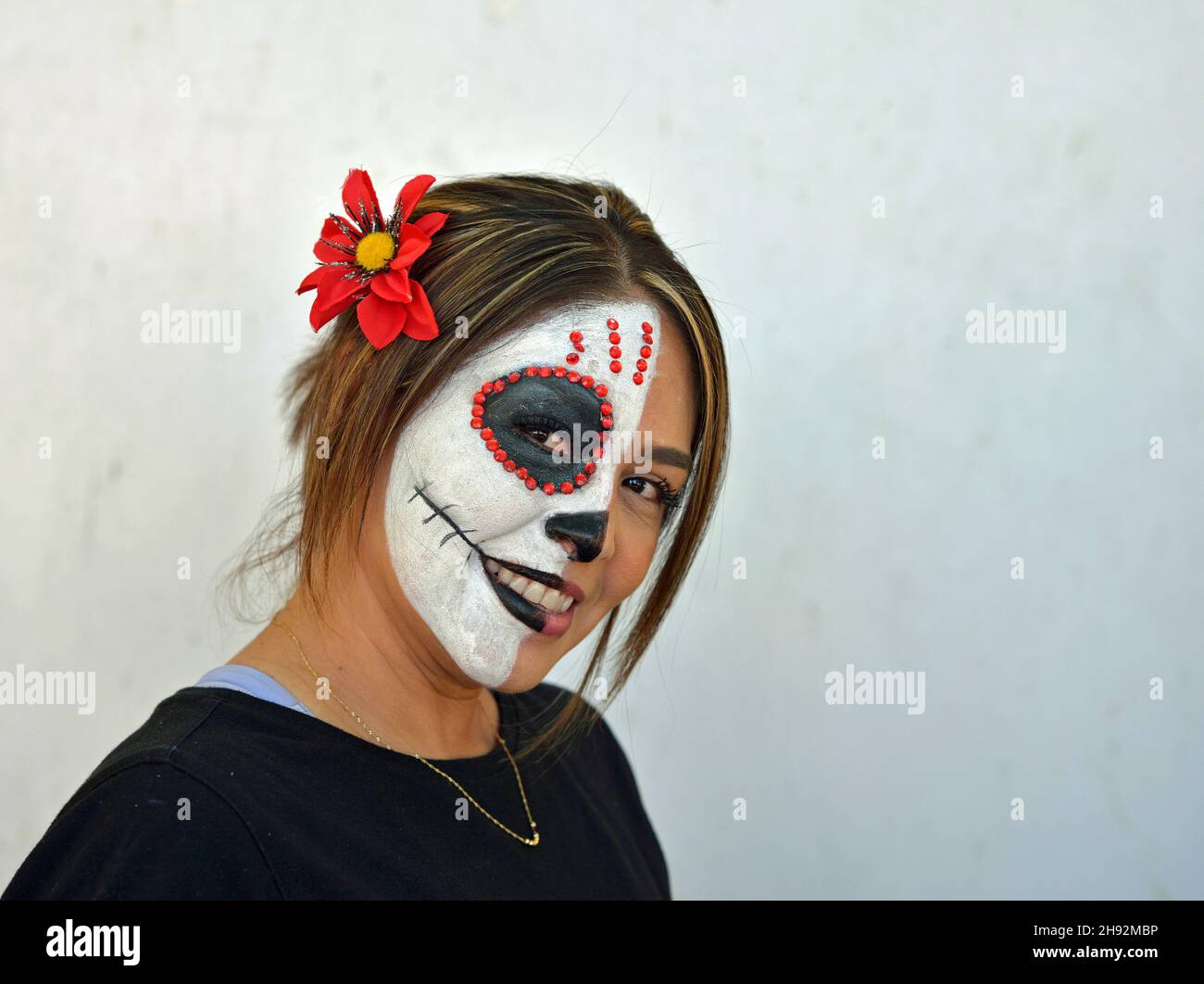 Die junge attraktive mexikanische Frau malt am Tag der Toten die rechte Hälfte ihres Gesichts mit einem schaurigen weißen Catrina Face Make-up und schaut den Betrachter an. Stockfoto