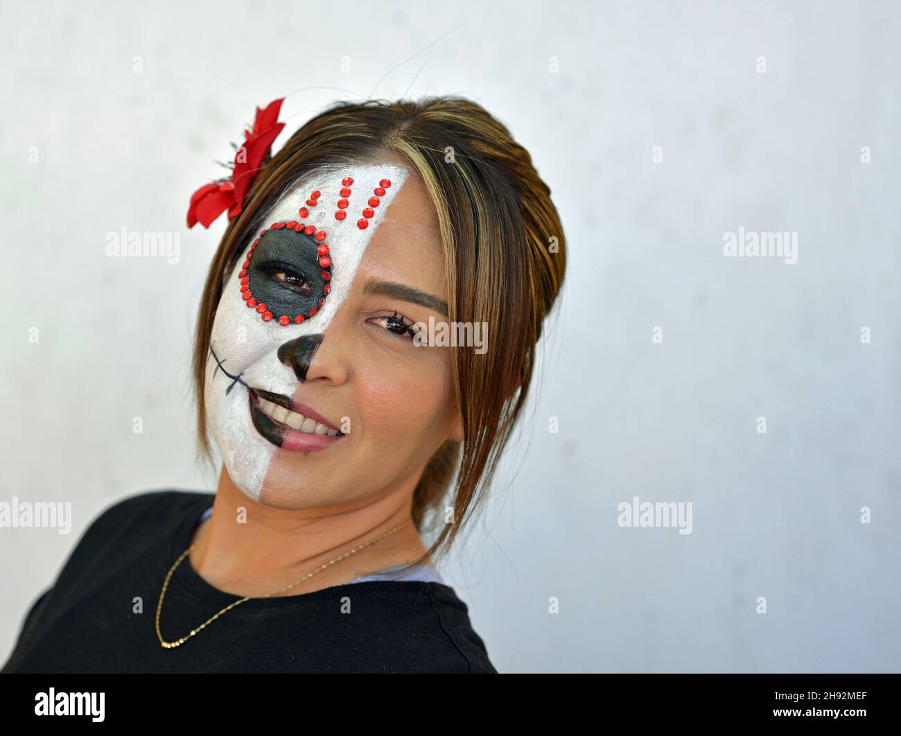 Die junge attraktive mexikanische Frau malt am Tag der Toten die rechte Hälfte ihres Gesichts mit einem schaurigen weißen Catrina Face Make-up und schaut den Betrachter an. Stockfoto