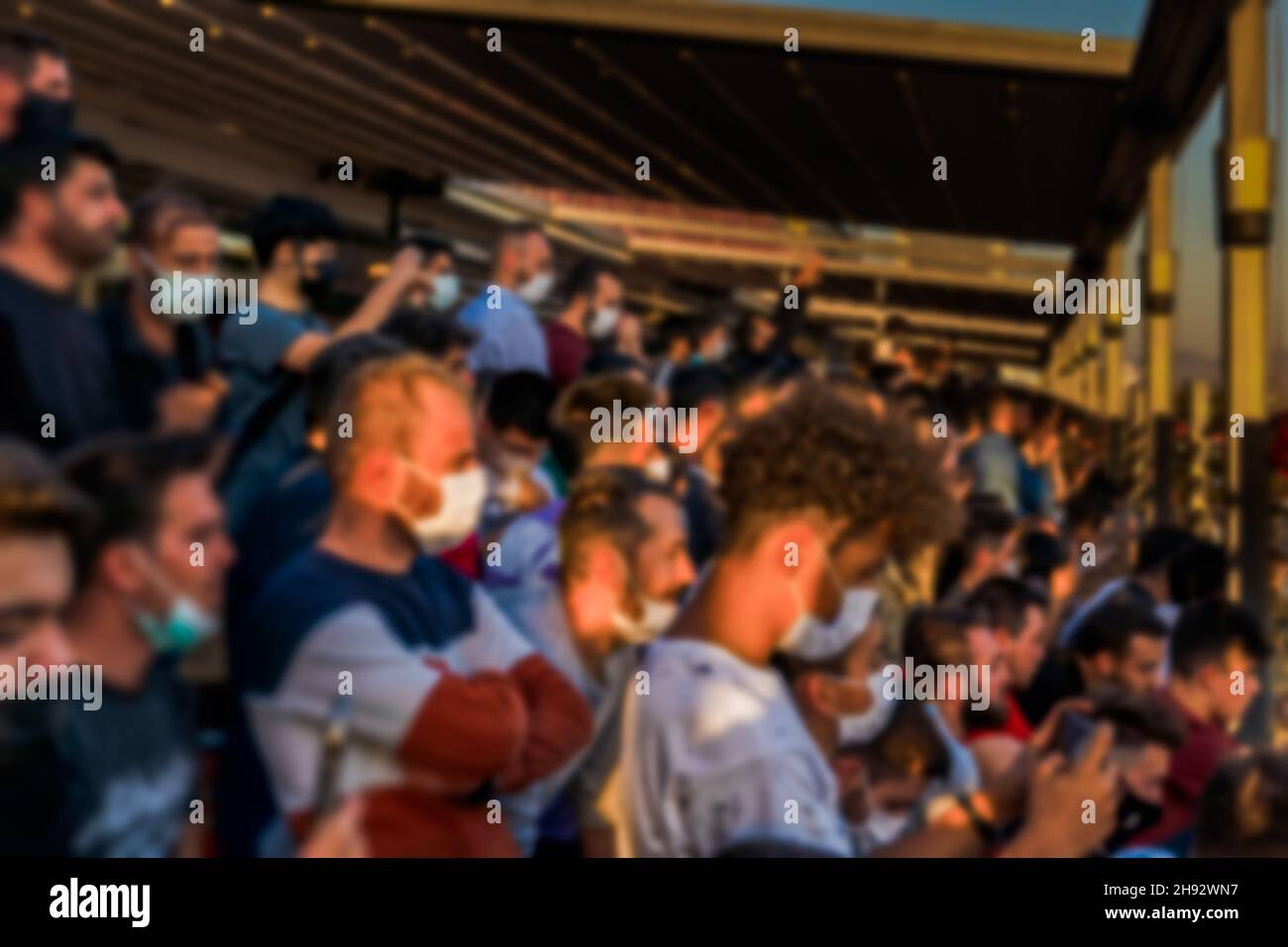 Unfokussiertes, unscharfes Gesicht maskierte Zuschauer, die das Spiel im Stadion mit einem Mobiltelefon in den Händen sehen. Stockfoto