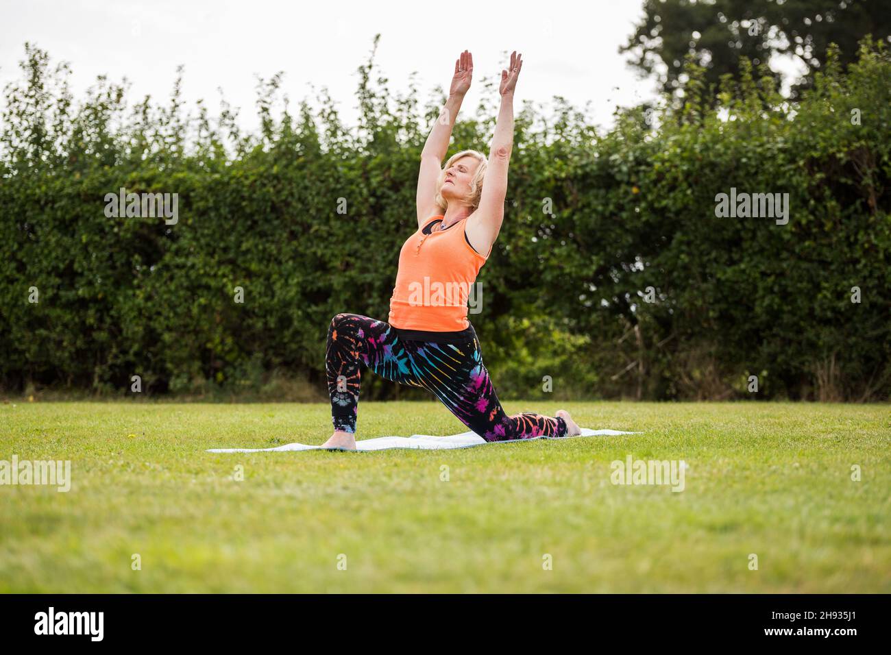 Eine Frau mittleren Alters Üben Yoga barfuß draussen in einem grünen Park. Sie trägt ein helles Orange Weste und schwarze Leggings. Der Stil des Yoga Sie i Stockfoto