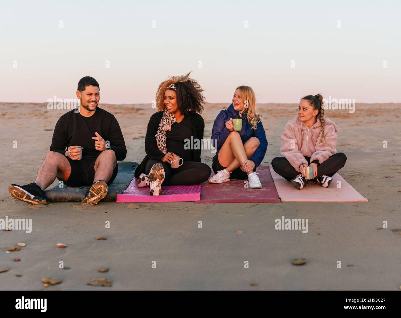 Multiethnische Gruppe von Menschen, die während des Sonnenuntergangs auf Matten mit Blick auf den Meereshorizont sitzen Stockfoto
