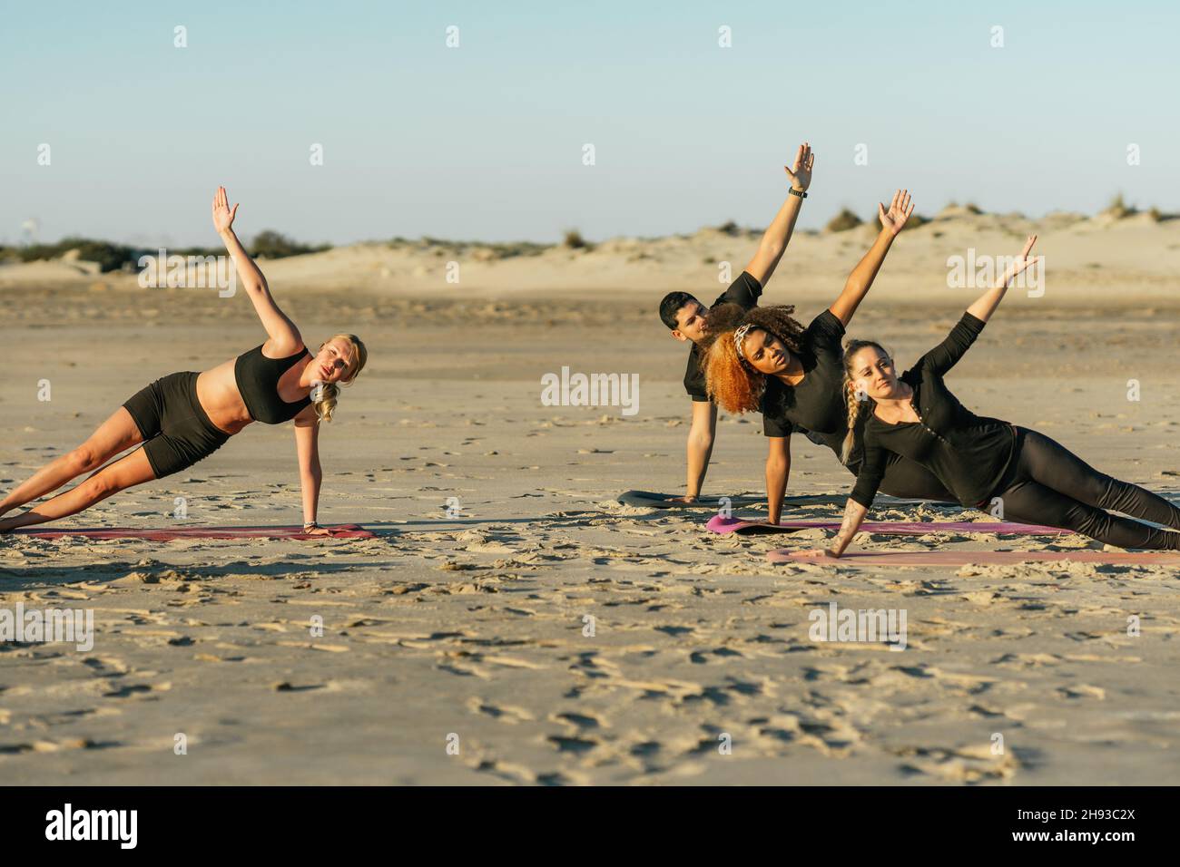 Yoga-Coach und eine Gruppe von multiethnischen Lernenden, die am Strand eine seitliche Planke machen Stockfoto