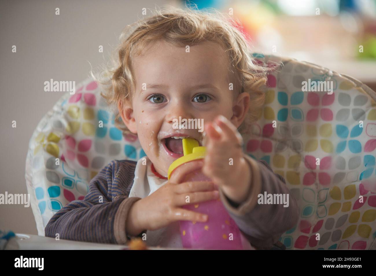 Ein 2-jähriges Mädchen lächelt, während sie auf einem Hochstuhl sitzt und mit einer Babyflasche spielt. Stockfoto