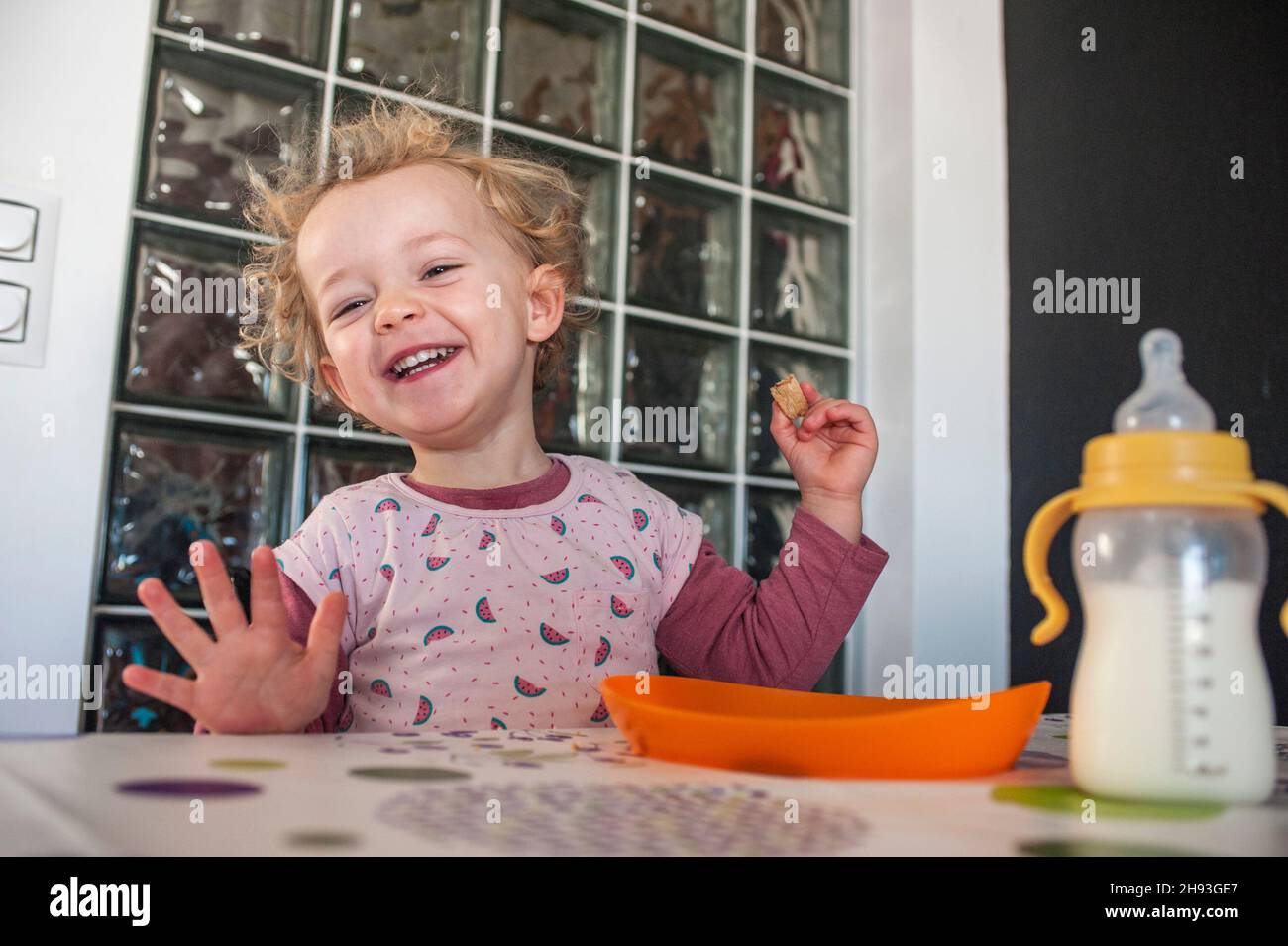 Ein 2-jähriges Mädchen lacht, als sie einen Snack und eine Babyflasche Milch hat. Stockfoto