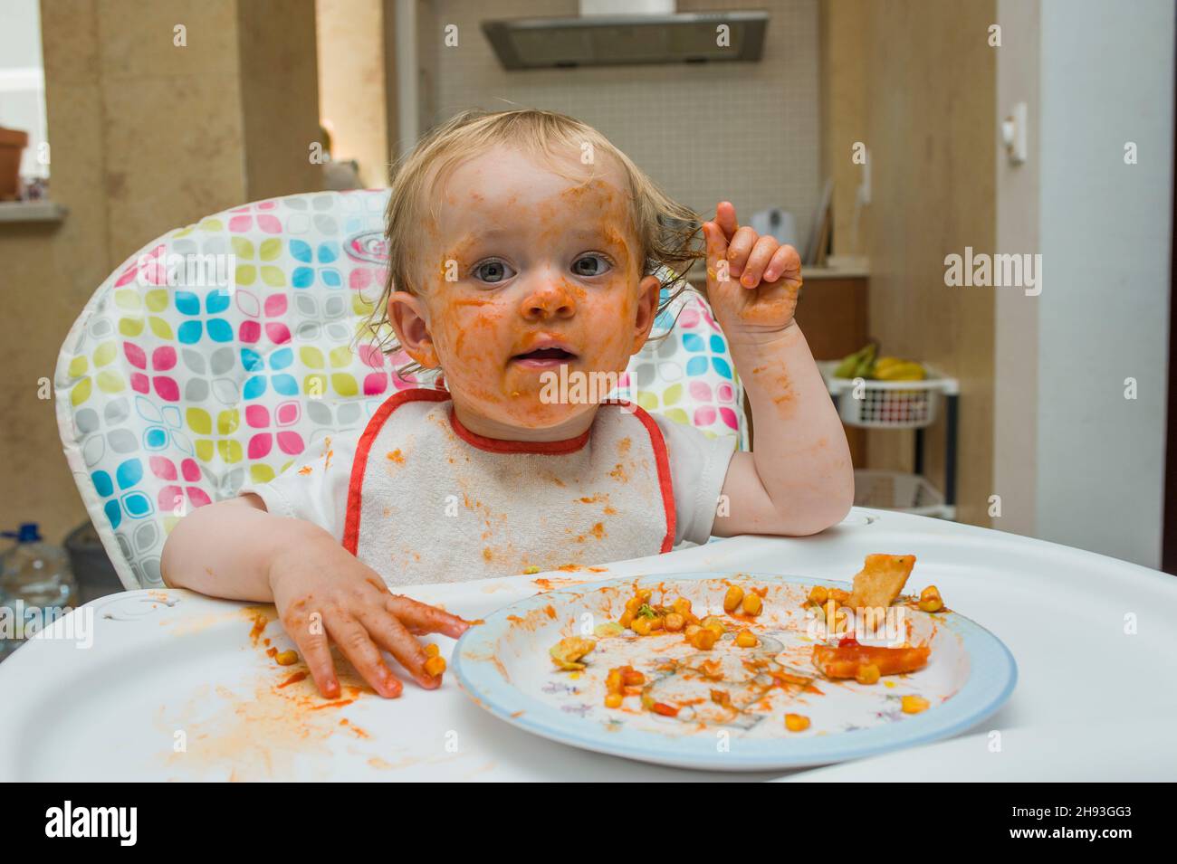 Ein kleines Mädchen (15 Monate alt) isst eine Mahlzeit aus süßem Mais und Tomatensauce auf unordentliche Weise und spielt mit ihrem Essen auf einem Hochstuhl in ihrem Haus. Stockfoto