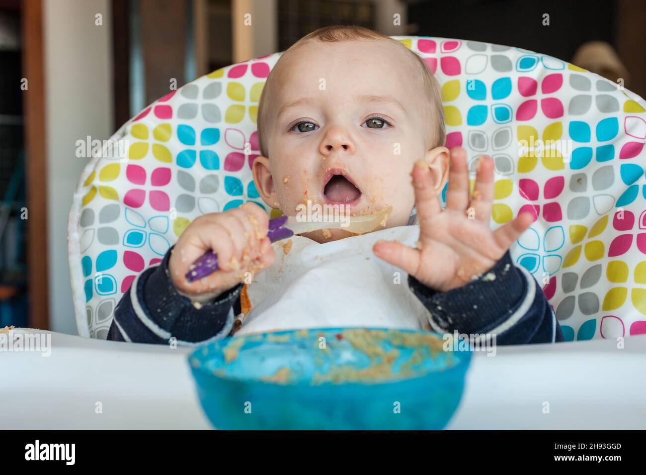 Ein kleines Mädchen (ca. 10 Monate alt) spielt mit ihrem Löffel, während sie ihr Abendessen mit pürierten Speisen zu sich nimmt. Stockfoto