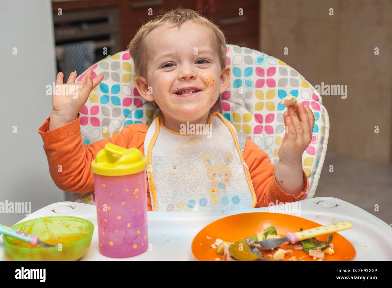 Ein 2-jähriges Mädchen lächelt, als sie auf einem Hochstuhl eine Mahlzeit isst. Stockfoto