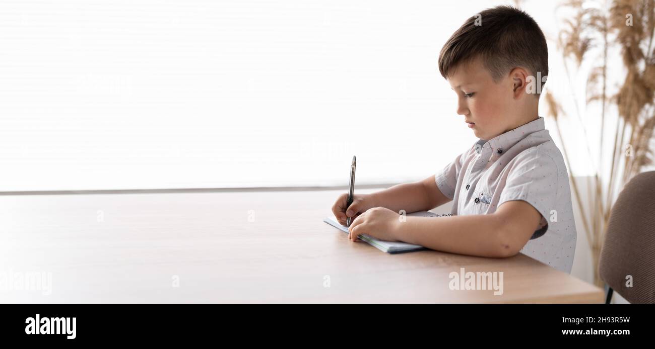 Konzentrierter Blick auf den Schuljungen am Schreibtisch, der am Fenster sitzt. Schüler schreibt Hausaufgaben in ein Notizbuch. Grundschüler, die den Test machen Stockfoto