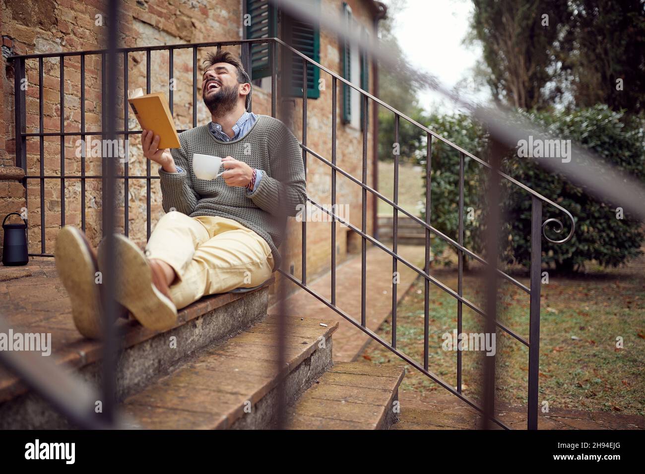 Ein junger Mann ist gut gelaunt, während er an einem schönen Tag auf der Veranda sitzt, warmen Tee trinkt und ein Interessantes Buch liest. Stadt, draußen, Ruhe Stockfoto