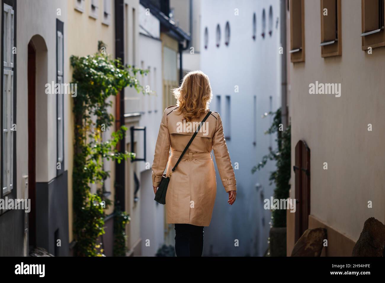 Stilvolle blonde Frau mit Trenchcoat und Spaziergang auf der Straße der Stadt. Modischer Trendstyle. Altstadtgebäude Stockfoto