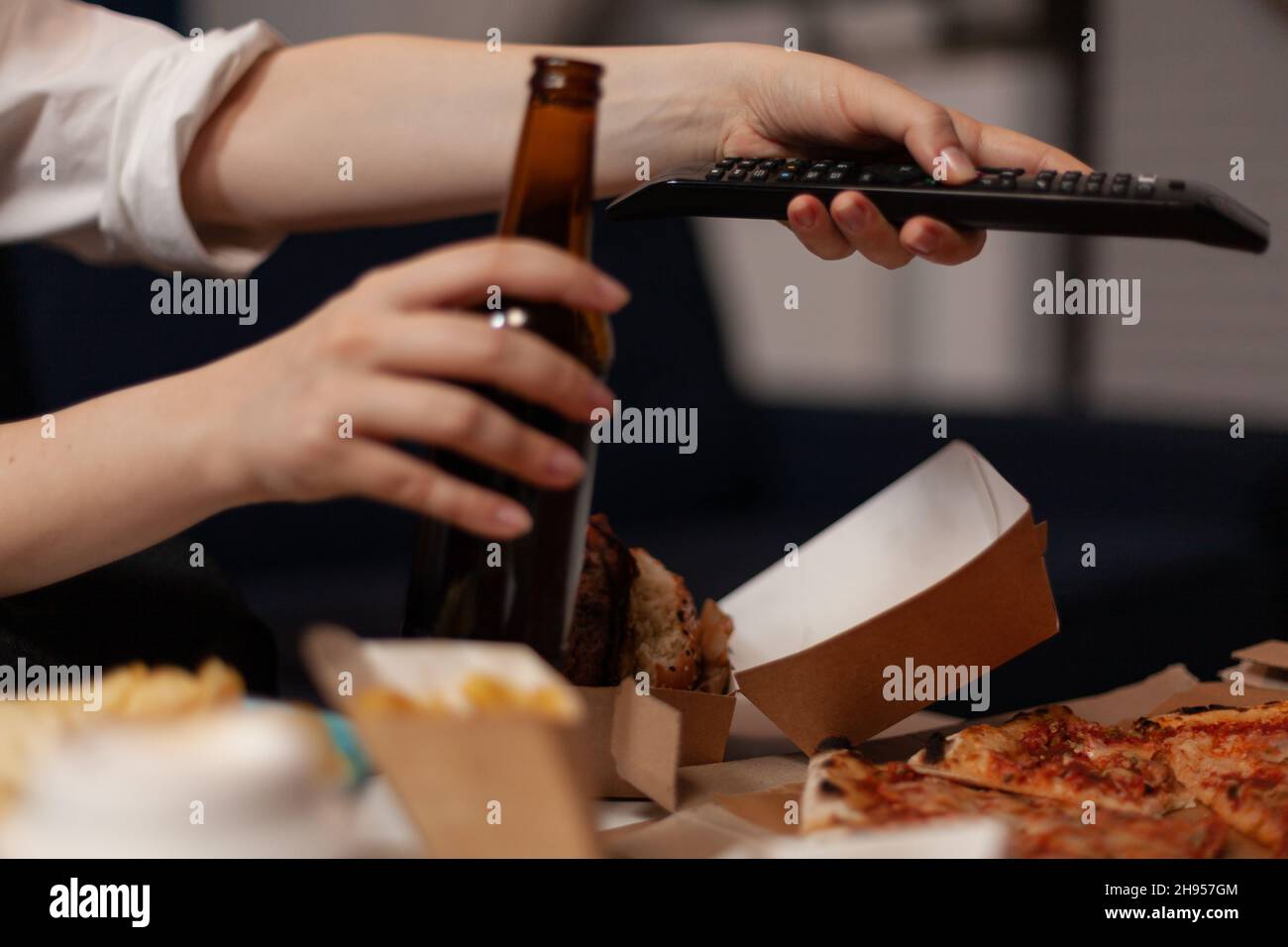 Nahaufnahme der Frau, die die Hände wechselt, mit der TV-Fernbedienung, die vor dem Tisch Fernsehen und Fast Food-Lieferservice ansieht. Kaukasische Frau hält Bierflasche und entspannt Pizza zum Mitnehmen. Stockfoto