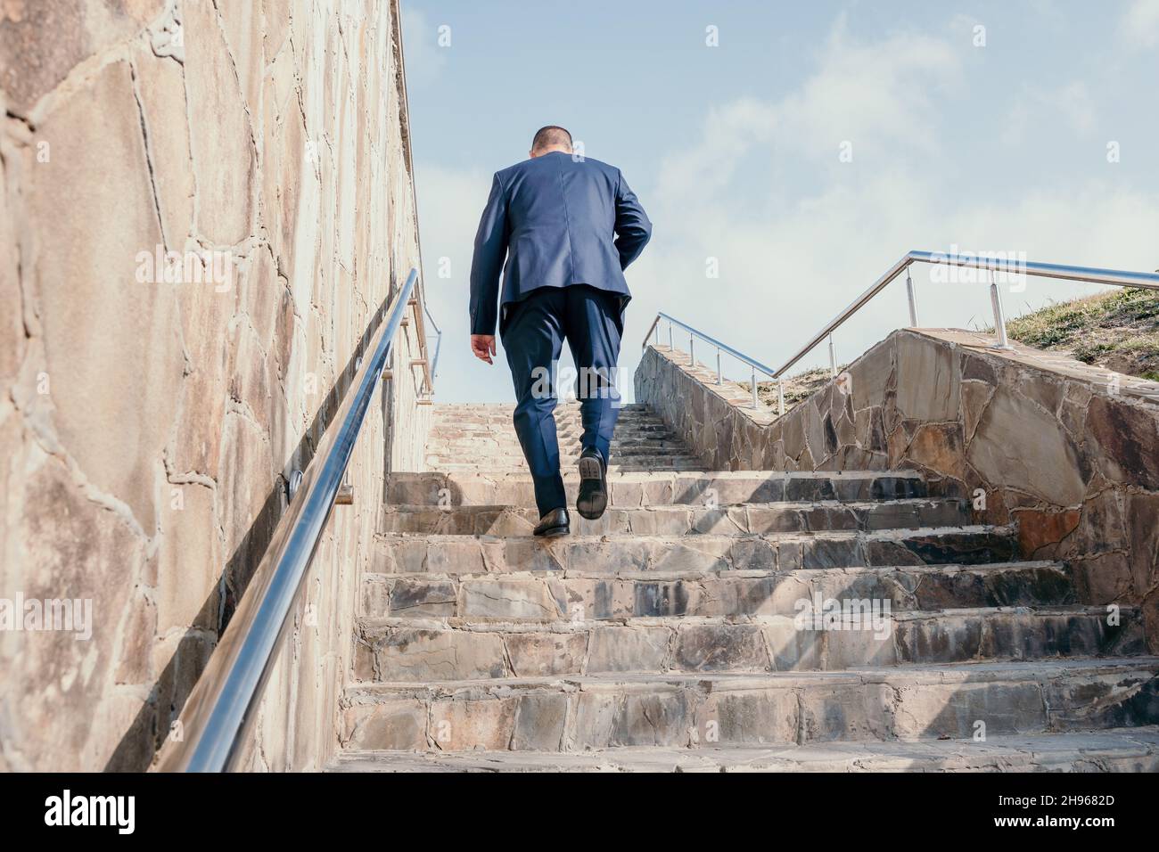 Selbstbewusster Geschäftsmann mittleren Alters mit Aktentasche, der die Treppe hoch läuft. Nahaufnahme eines Geschäftsmannes in einem blauen Anzug mit Tragetasche und einer Kaffeetasse, der die aufsteigen kann Stockfoto