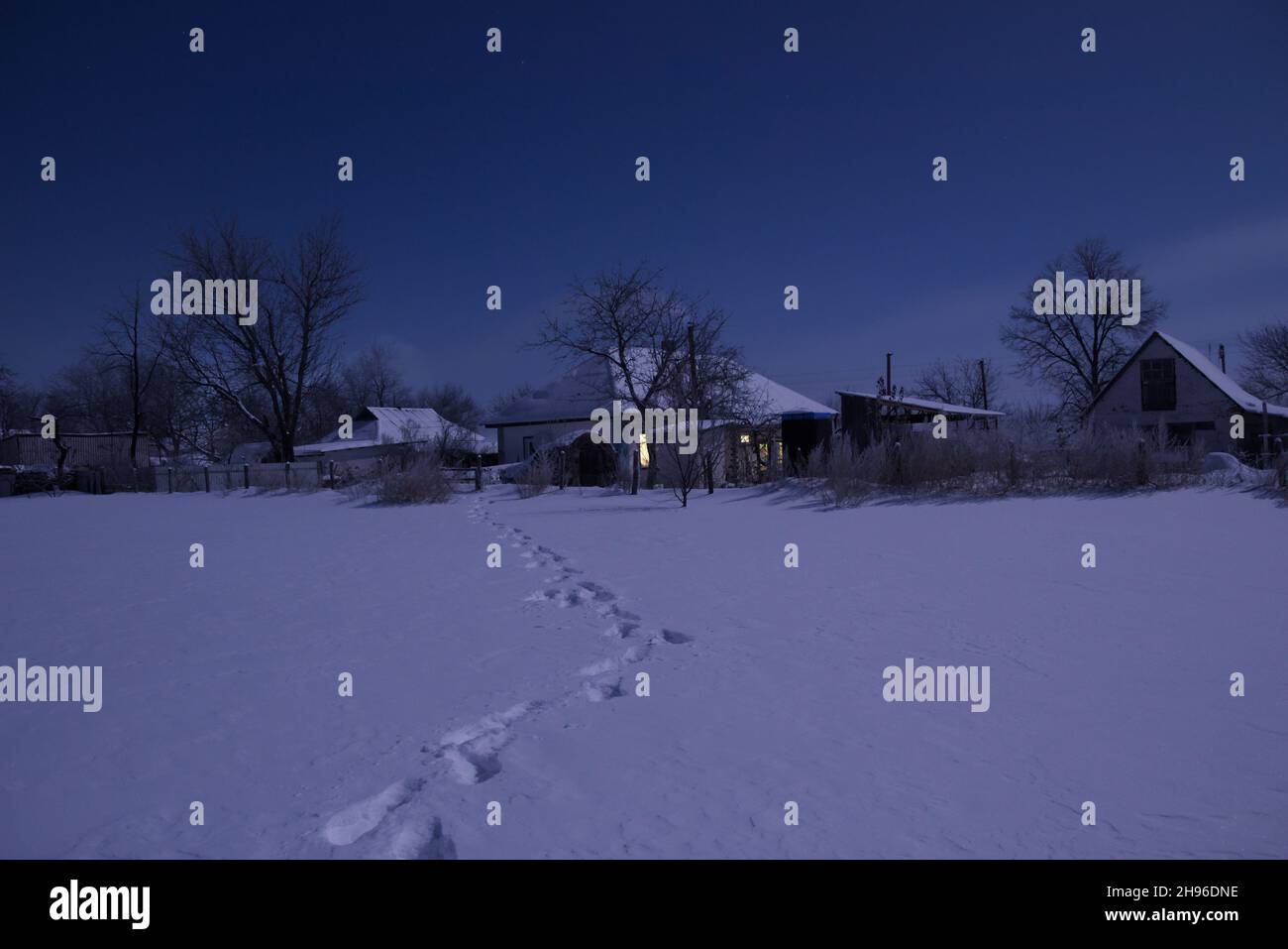 Nachtlandschaft eines schneebedeckten Feldes. In der Ferne sieht man das Dorf. Es gibt die Fußabdrücke auf dem Schnee. Stockfoto