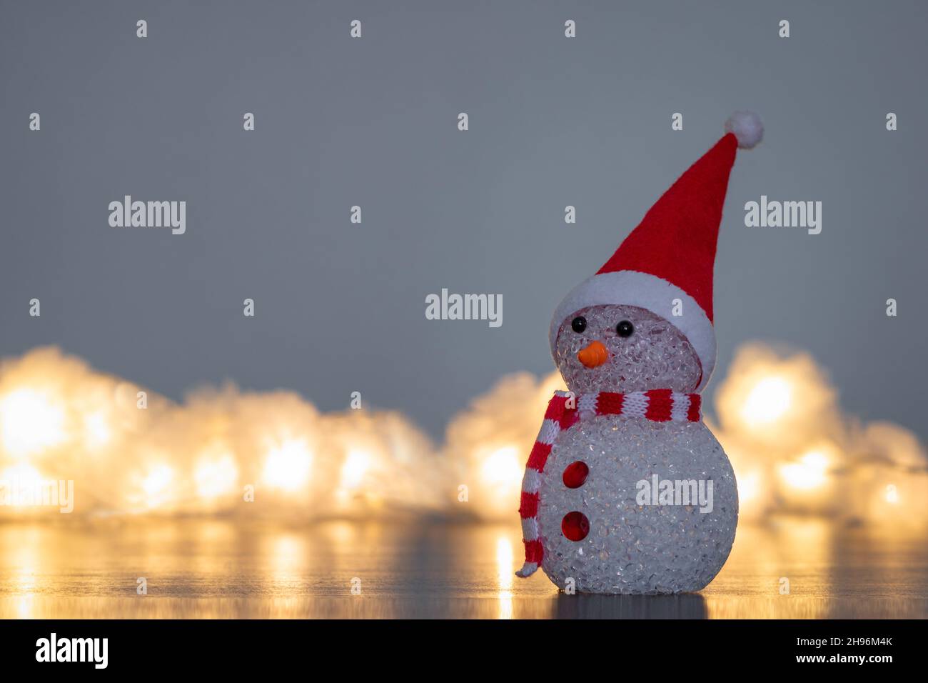 Kleine Figur eines niedlichen Weihnachtsschneemanns, der einen rot-weißen Schal und einen Weihnachtsmann-Hut mit verschwommenen Lichterketten trägt Stockfoto