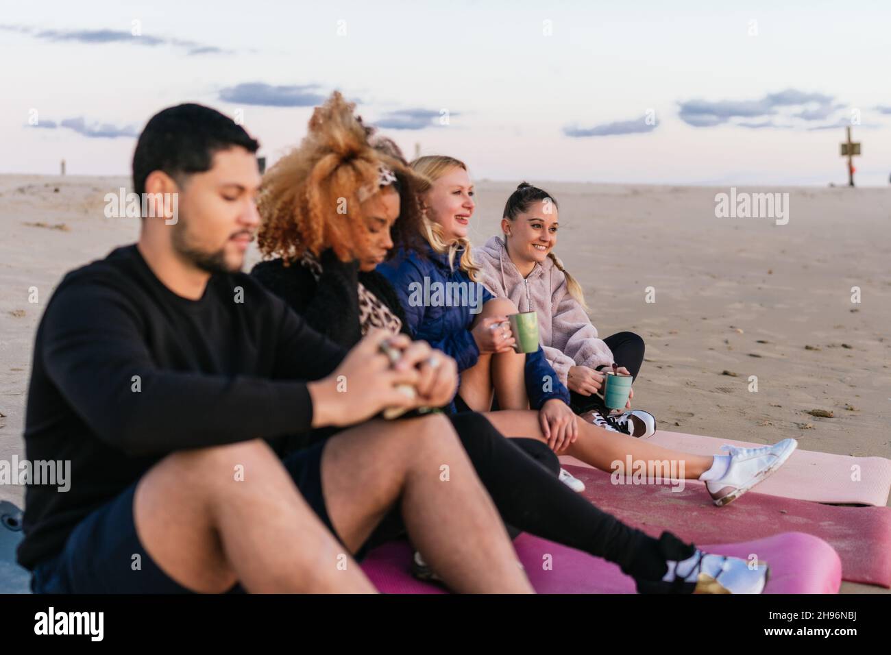 Multikulturelle Gruppe von Menschen, die am Strand sitzen und während des Sonnenuntergangs gemeinsam Tee trinken Stockfoto