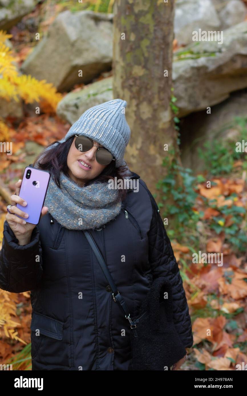 Porträt einer Frau mit Sonnenbrille und Wollhut, die an einem Herbsttag morgens mit dem Smartphone im Wald Selbstporträts aufnahm. Stockfoto