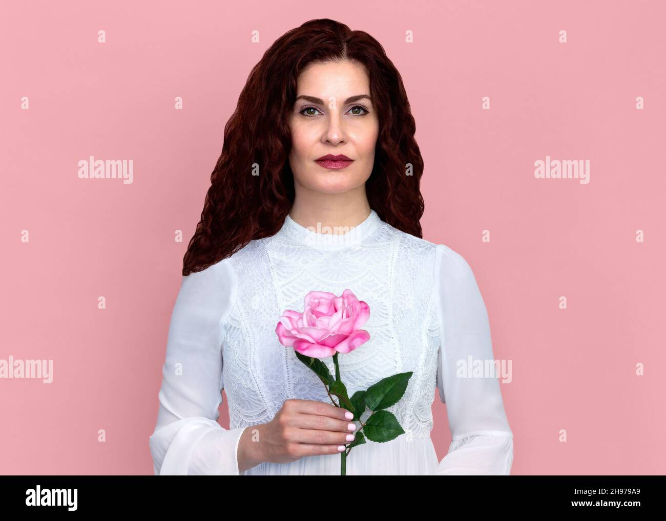 Frau mit einer Rose gegen rosa Hintergrund. Nahaufnahme Porträt der schönen Frau mit welligen rotbraunen Haaren hält einzelne rosa Rose zart. Stockfoto