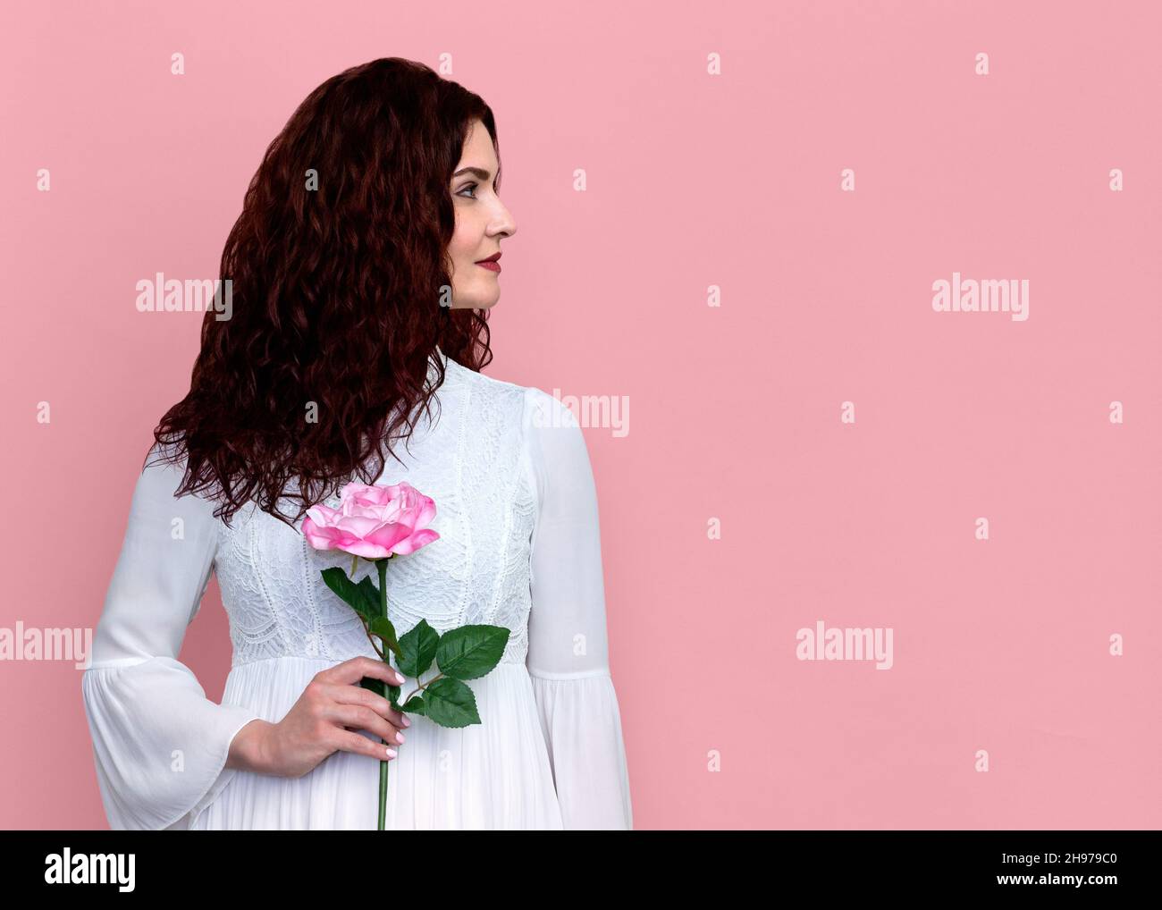 Frau mit einer Rose, die zur Seite auf einem rosa Hintergrund schaut. Porträt einer hübschen Frau mit einer rosa Rose in der Hand. Platz für Kopien. Stockfoto