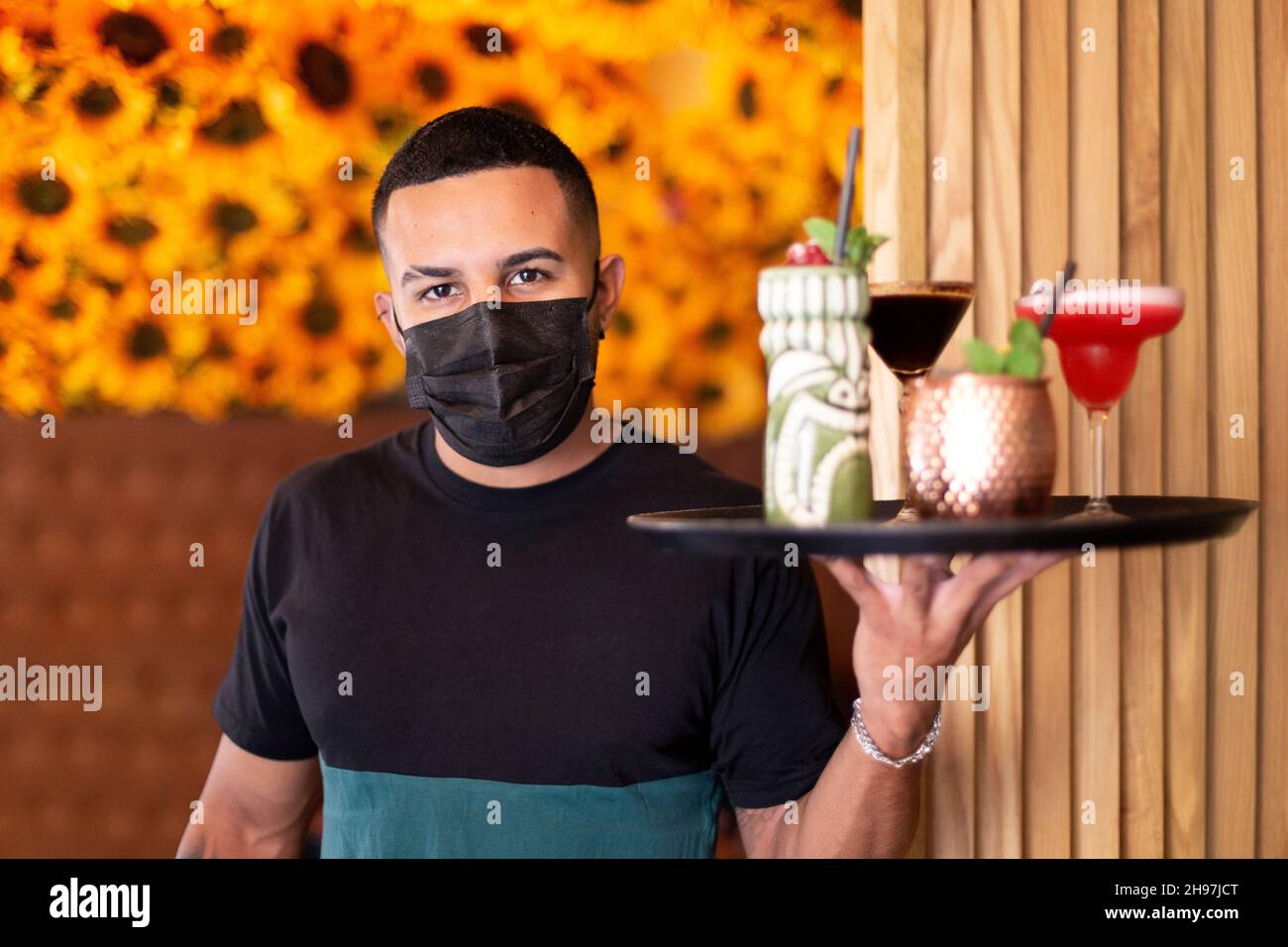 Ein junger lateinamerikanischer Kellner serviert Cocktails auf einem Tablett. Er trägt eine medizinische Maske während der Arbeit. Stockfoto