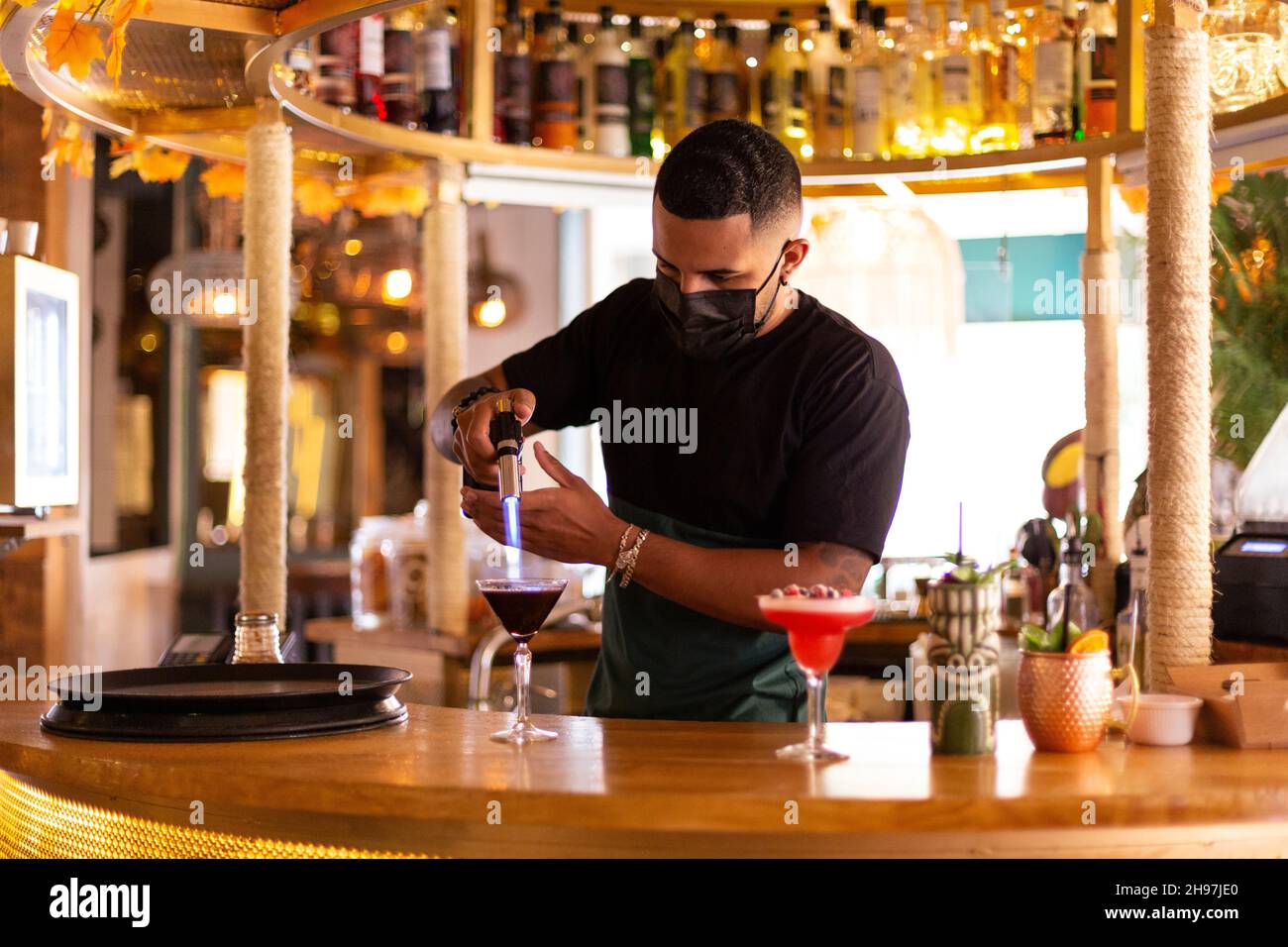 Lateinamerikanischer Barkeeper mit Gesichtsmaske hinter der Bar in einer modernen Cocktailbar. Platz für Text. Stockfoto