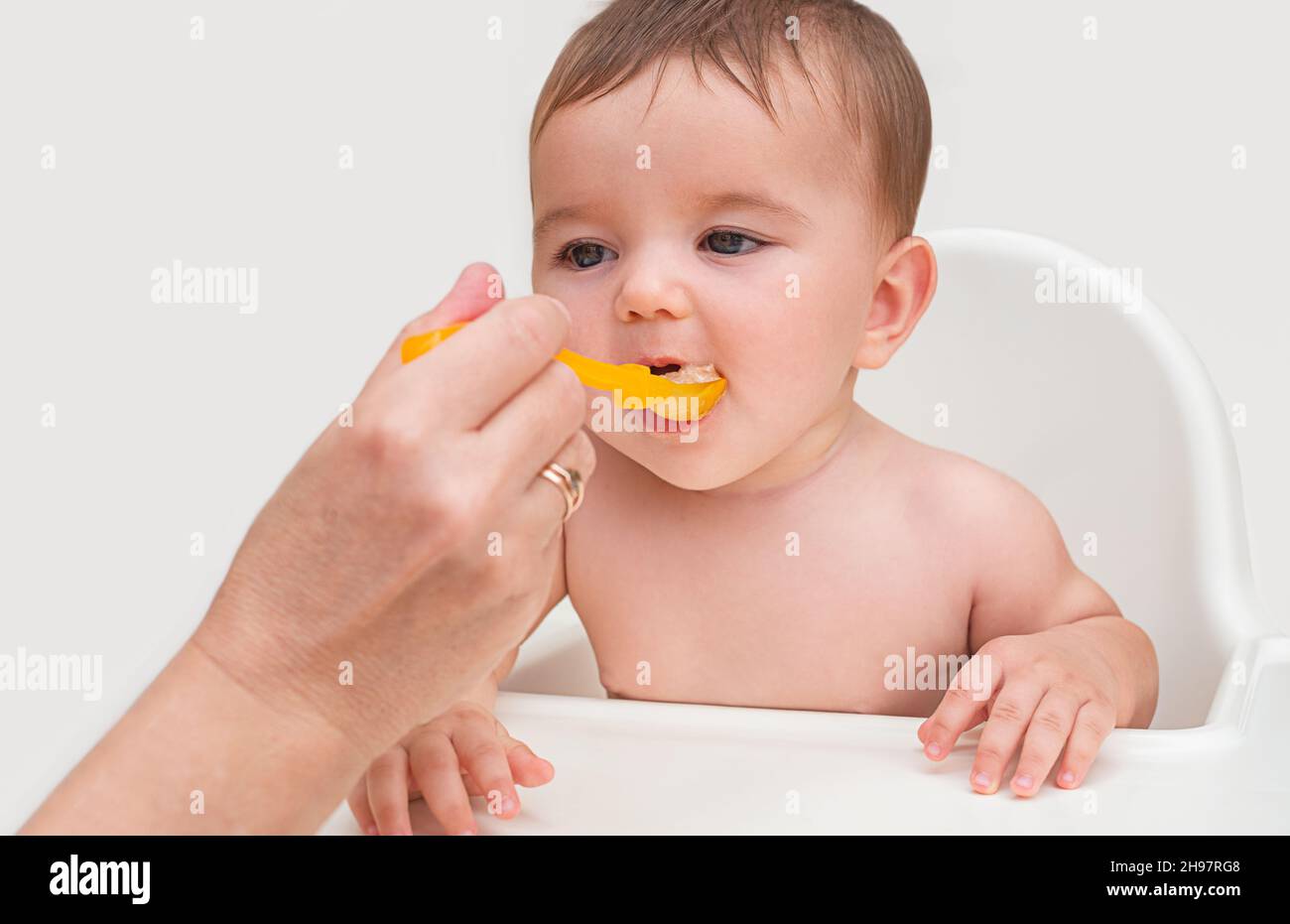 Die erste Fütterung des Babys. Das Kind öffnet seinen Mund, um natürliches Orangenpüree aus einem orangefarbenen Löffel aus der Hand seiner Mutter zu essen Stockfoto