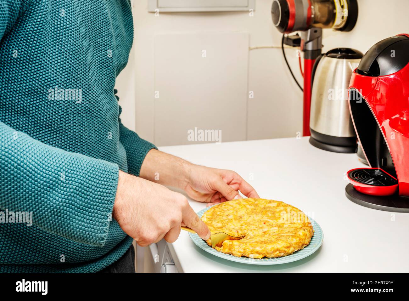 Die Hand eines jungen Mannes hält eine goldene Gabel, um ein frisch zubereitetes spanisches Omelett auf einer weißen Küchentheke zu essen Stockfoto