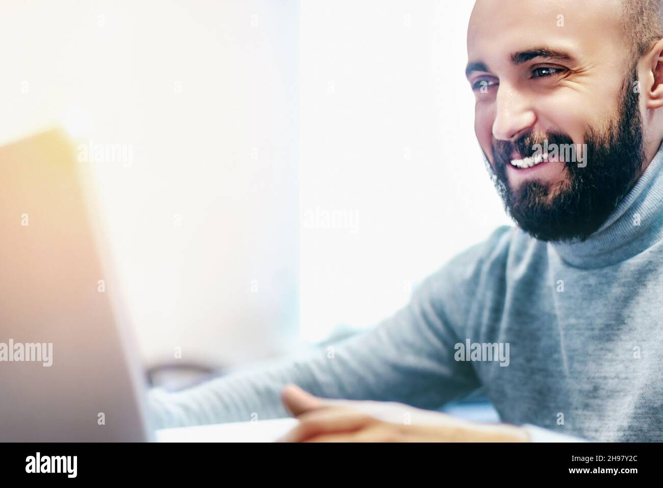 Junger Mann mit Bart blickt auf den Laptop-Bildschirm und lächelt. Internet-Dating oder Kommunikation durch Videokommunikation. Junger positiver Mann mit arabischem Aussehen. Emotionales Nahaufnahme-Porträt. Stockfoto