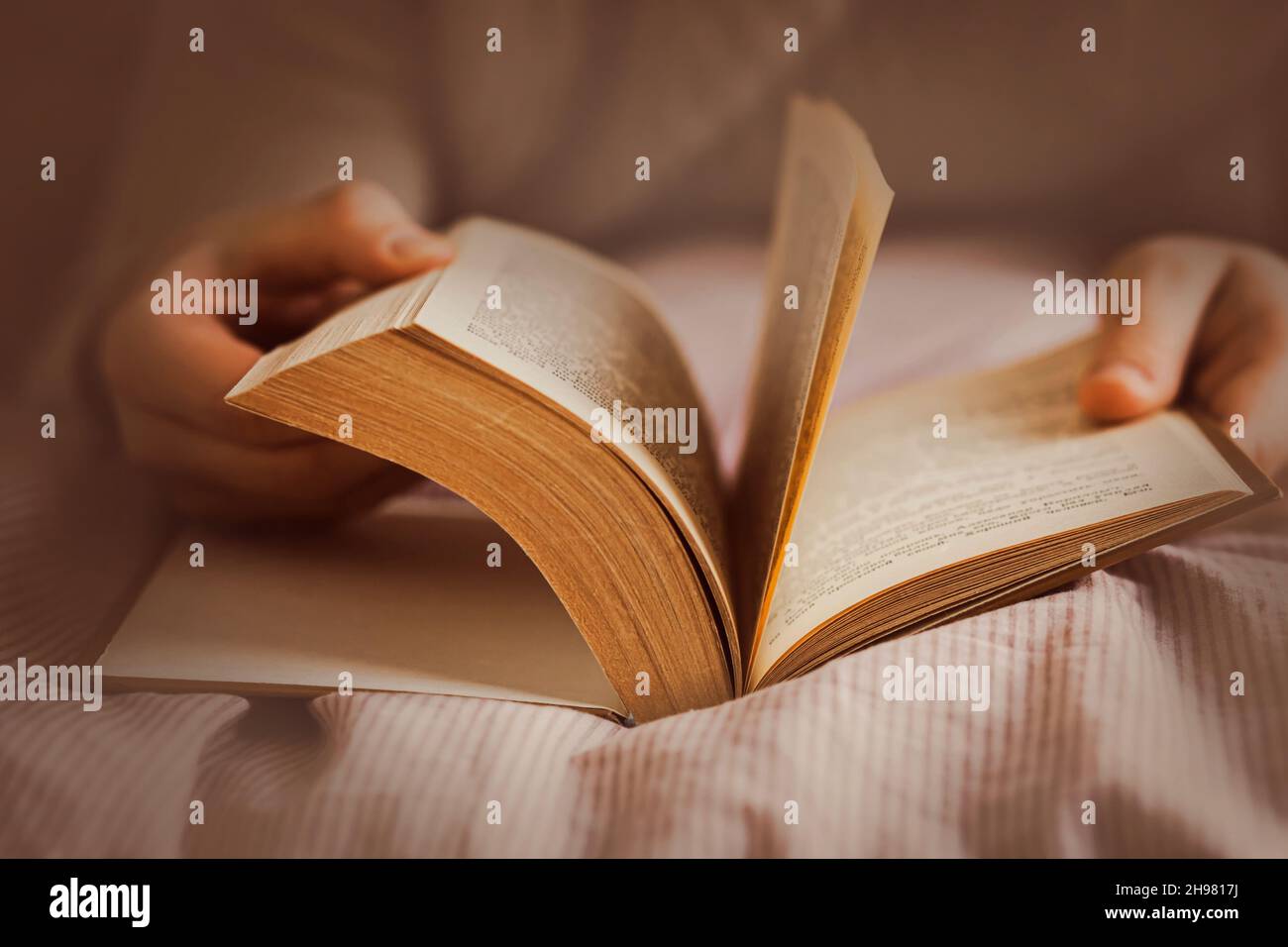 Ein Mann, der auf einem weichen Bett mit einer gestreiften Decke liegt, blättert durch ein dickes altes Buch. Entspannen beim Lesen eines interessanten Romans. Selbstentwicklung. Stockfoto