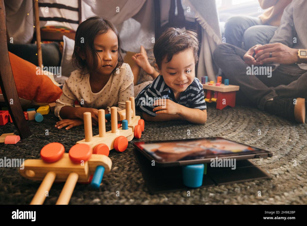 Junge Geschwister sehen sich Inhalte von Kindern auf einem digitalen Tablet an. Zwei entzückende Kleinkinder liegen auf dem Boden, ihre Eltern sitzen im Hintergrund. Sibli Stockfoto