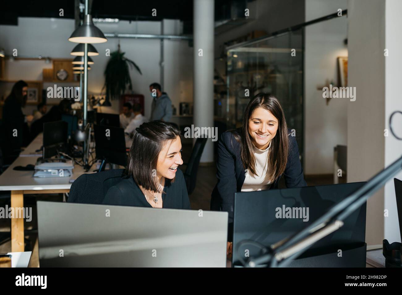 Lächelnde Geschäftsfrau, die einem Kollegen zuhört, vor einem Computer, am Schreibtisch und im Büro. Sie sind in klugen Casuals. Stockfoto