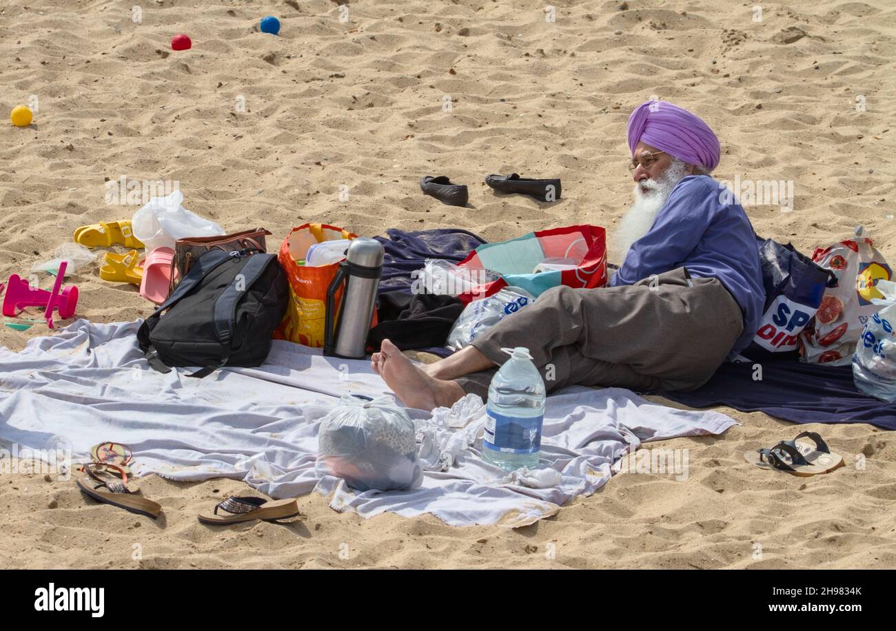 Sikh man in a Turban Laying on Bournemouth Beach in the Sun Umgeben von Einem Picknick, Beachwear und Games, Boscombe UK Stockfoto