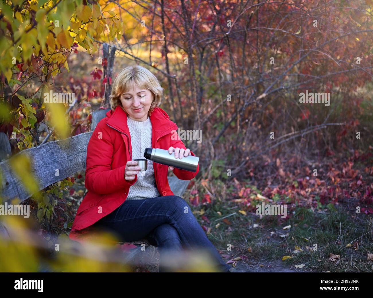 Eine angenehme Frau von 50-60 Jahren in einem Herbstpark, die auf einer Holzbank sitzt, gießt Tee aus einer Thermoskanne, um sich warm zu halten. Stockfoto