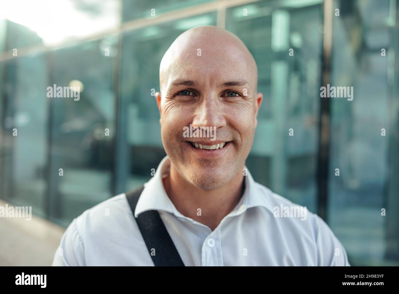 Fröhlicher Geschäftsmann, der fröhlich vor der Kamera lächelt, während er vor einem Bürohochhaus in der Stadt steht. Erfolgreiche Unternehmungskrisen für mittlere Erwachsene Stockfoto