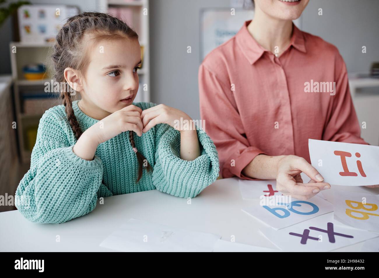 Kleines Mädchen, das am Tisch sitzt und der Lehrerin zuhört, die ihr während einer Lektion das englische Alphabet zeigt Stockfoto