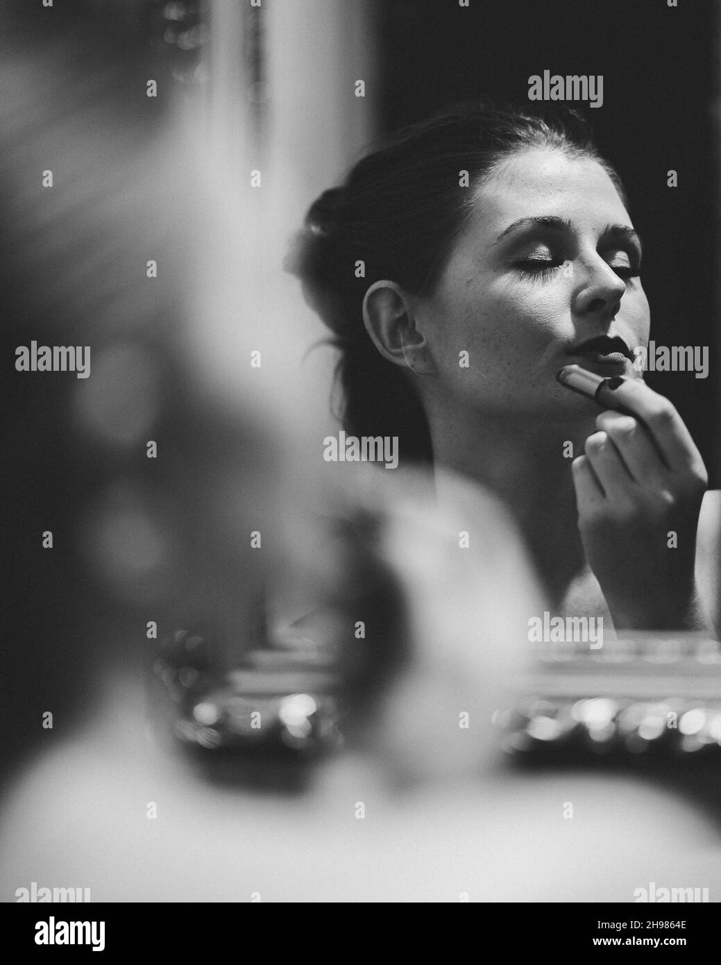 Ein graues Porträt einer jungen Brünette, die Lippenstift vor dem Spiegel aufsetzt Stockfoto